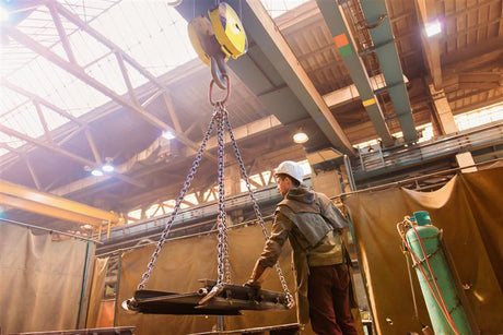 Factory worker wearing a hard hat and safety gear operating an overhead crane with chains inside an industrial warehouse.